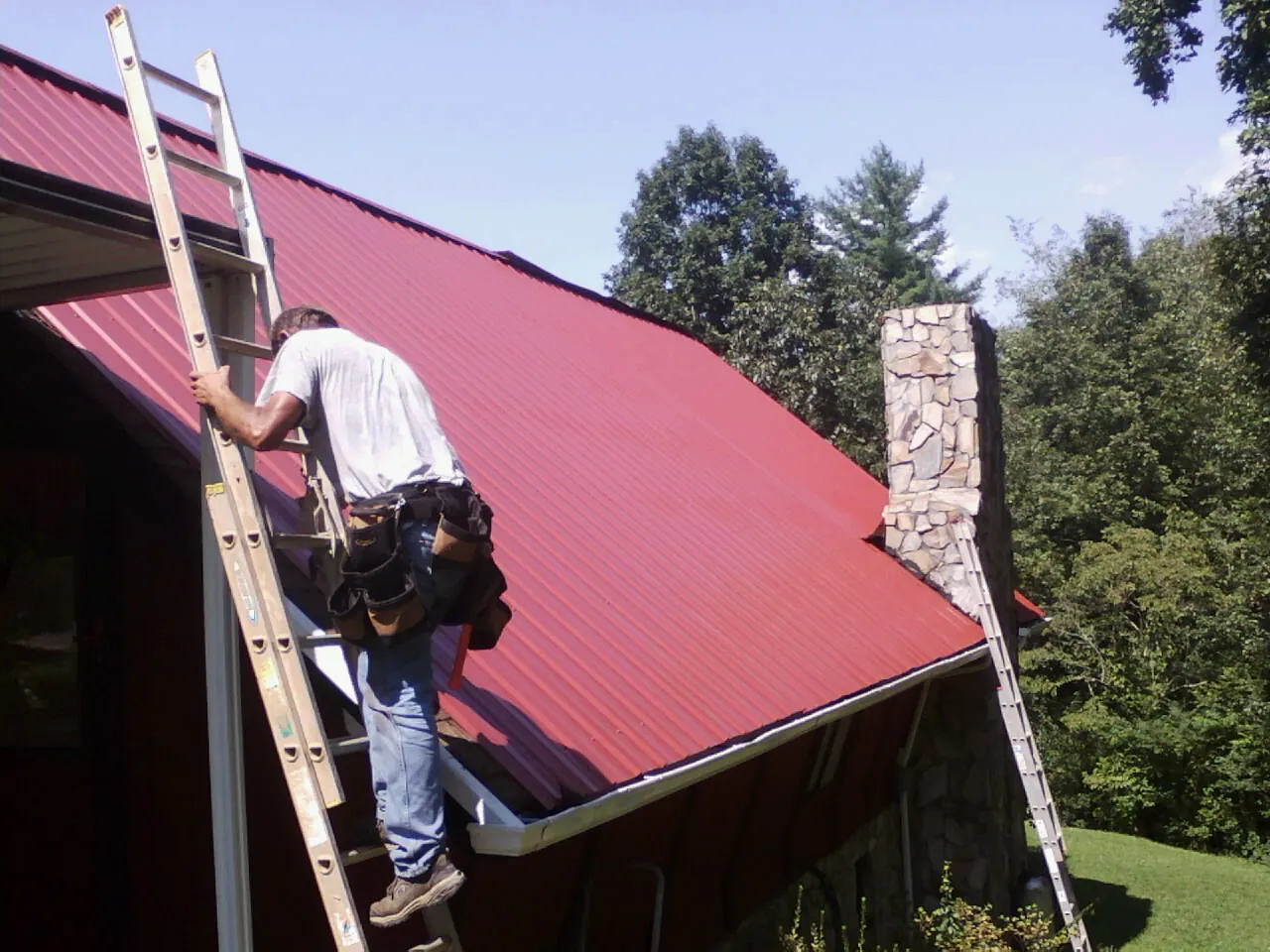 Roofer on a ladder beside a red metal roof and stone chimney — Soffit & Fascia Repair by Roofing Brothers in South Plymouth